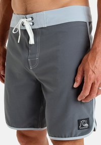 Gray swim shorts with a light gray waistband, white drawstring, and contrasting white stitching. Features a side pocket and a logo patch.
