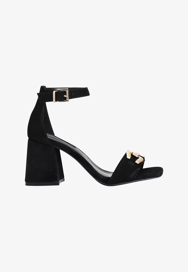 Sandalias de tacón alto de ante negro con punta cuadrada y un tacón grueso. Presentan una correa en el tobillo con una hebilla metálica y un acento decorativo dorado.