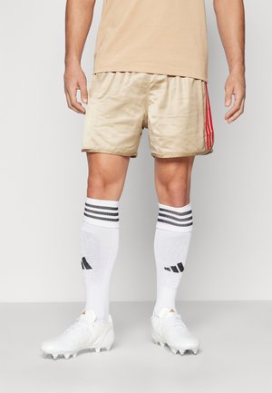 Beige Sportshorts mit vertikalen Nadelstreifen und roten Seitenstreifen, kombiniert mit weißen knielangen Socken mit schwarzen Streifen und weißen Fußballschuhen.