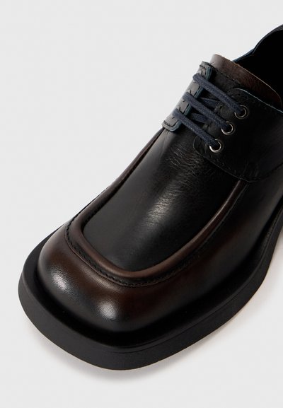 Black leather shoe with a rounded toe, featuring brown accents, dark blue laces, and a textured finish on the upper, mounted on a sturdy sole.