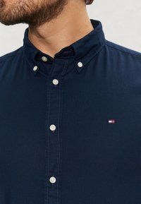 Camicia button-up in cotone blu navy con colletto, dotata di bottoni chiari e un piccolo logo in rosso, bianco e blu vicino al petto.
