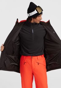 Een heren ski-outfit met een zwarte tussenlaag en een zwarte buitenjas met rode accenten, gecombineerd met feloranje broeken en een skibril.