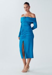 Robe bleue à épaules dénudées avec une texture froncée, manches longues, fente frontale et détail de fermeture éclair centrale. Associée à des talons dorés à brides.