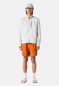 Hellgraue Kapuzenjacke mit Reißverschluss, dekorativen Nähten und einer Brusttasche; kombiniert mit orangenen Shorts und beigen Sandalen.