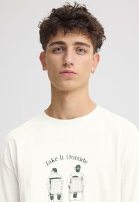 T-shirt en coton blanc avec un imprimé vert représentant deux personnes assises sur des chaises pliantes, avec le texte "Prenez l'air" au-dessus.