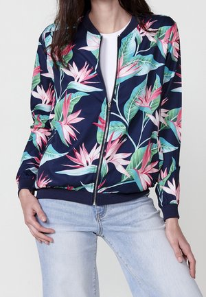 Vrouw draagt een marineblauwe jas met rits, voorzien van een roze en groene tropische bloemenprint, gecombineerd met lichtblauwe spijkerbroek en een witte top.