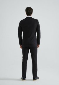Costume noir avec veste et pantalon ajustés, tissu lisse, peu de détails. Porté avec des chaussures sombres, vu de dos sur un fond uni.