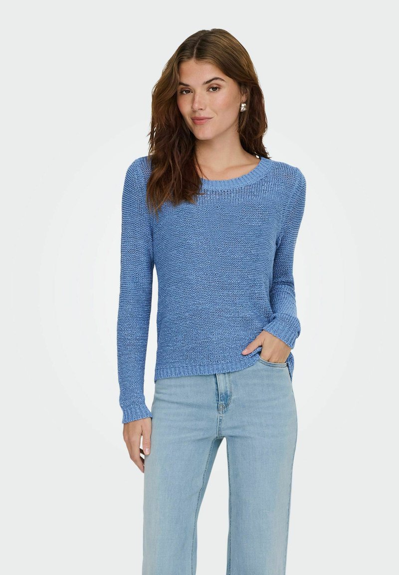 Jeune femme aux longs cheveux bruns portant un pull en tricot bleu et un jean bleu clair, posant avec une main dans la poche devant un fond uni.