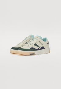 Sneakers featuring a beige leather upper, light blue accents, dark green and navy detailing, perforated texture, and a white rubber sole.