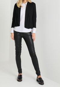 Svart kort cardigan med långa ärmar över en vit skjorta med knappar, i kombination med svarta byxor i konstläder och svarta loafers med fransar.