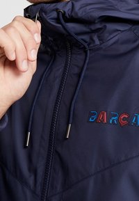 Marineblauwe hooded jas met een ritssluiting, voorzien van een trekkoord, een gladde textuur en een meerkleurig "BARÇA" logo op de borst.