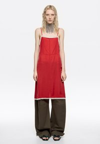 Kobieta-modelka o falowanych blond włosach ubrana jest w czerwoną sukienkę na ramiączkach nałożoną na szerokie oliwkowe spodnie oraz srebrny naszyjnik z frędzlami, stojąc na białym tle.