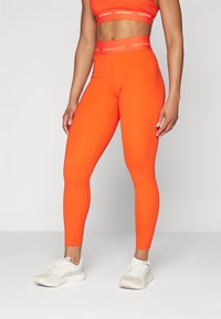 Ryškiai oranžinės aukštos liemens sportinės leggingės su lygia tekstūra ir "New Balance" logotipu ant juosmens, derinamos su šviesos spalvos bėgimo bateliais.