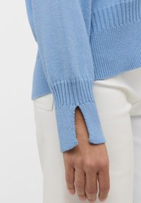 Maglione azzurro chiaro in maglia con polsini a costine e uno spacco laterale. La trama del tessuto è liscia e morbida, abbinata a pantaloni bianchi.