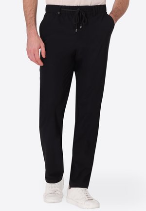 Uomo che indossa pantaloni neri con coulisse, con le mani nelle tasche e sneakers bianche, in piedi davanti a uno sfondo uniforme.