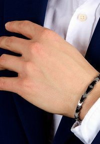 Silver metal bracelet with interlocking links, featuring a subtle engraved detail. Worn on a hand against a navy suit jacket.