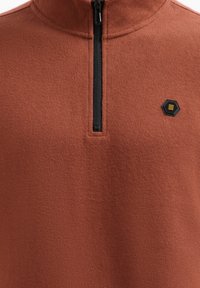 Rits-fleece pullover in roestkleur, met een textielstof, hoge kraag en zwarte rits. Klein zwart hexagonaal logo op de borst.