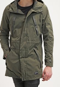 Parka verde oliva con cappuccio, chiusura frontale con zip, due tasche sul petto e tasche laterali. Realizzata in materiale leggero, presenta chiusure a scatto e a zip.