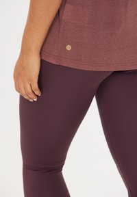 Leggings deportivos en color burdeos con una textura suave y un diseño ajustado, combinados con una camiseta de manga corta morada que presenta un detalle del logo en dorado.