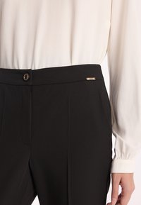 Czarne spodnie z dopasowanym stanem, z złotym guzikiem i małym akcentem w postaci logo. Materiał jest gładki z przodu z plisami.