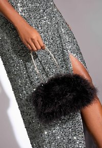 Juoda dirbtinė kailio rankinė su trumpomis rankenomis, laikoma rankoje; dėvima su sidabrine blizgučių suknele, turinčia aukštą iškirpimą.