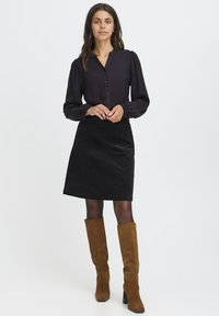Juoda marškinėlių blusa su pūstomis rankovėmis, derinama su juoda kordurojo sijonu. Rudos veliūrinės į kelią siekiančios botai užbaigia aprangą.