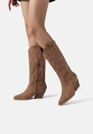 Stivali da cowboy color cuoio al ginocchio in pelle scamosciata con motivi traforati, chiusure laterali con cerniera e tacchi a blocco indossati su gambe nude su sfondo bianco.