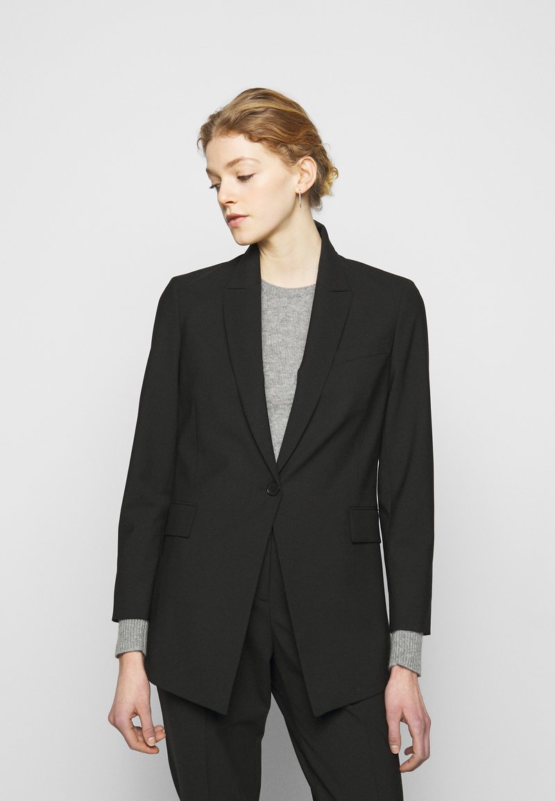 Blazer preto sob medida com um único botão, lapelas recortadas e dois bolsos frontais. Usado sobre um suéter cinza, apresentando um tecido liso e um caimento estruturado.