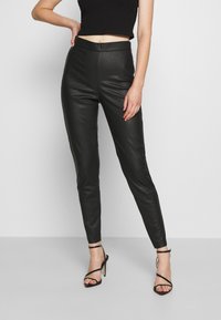 Czarne skórzane legginsy z wysokim stanem, gładką fakturą i wyraźnymi szwami, zestawione z czarnymi sandałami na wysokim obcasie z paskami.