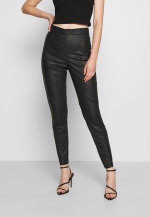 Pantalons en cuir noir ajustés avec une taille haute, texture lisse et coutures profilées, associés à des sandales à talons hauts noires à lanières.
