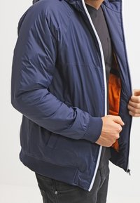 Giacca a vento imbottita blu navy con cappuccio, dotata di cerniera bianca, polsini elastici e fodera arancione brillante; materiale liscio e resistente all'acqua.