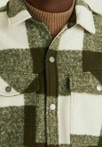 Veste en laine verte et crème à carreaux avec boutons-pression et deux poches à rabat sur le devant, portée sur un pull à col roulé côtelé marron.