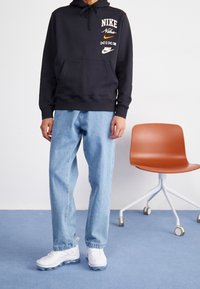 Svart Nike-hoodie med vita och orange detaljer, blå denimjeans och vita träningsskor med klara detaljer, står på blå matta.