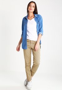 Jonge vrouw staat, gekleed in een blauwe denim shirt over een witte T-shirt, beige broek en witte sneakers tegen een plain witte achtergrond.