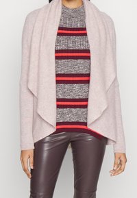 Helles rosa Cardigan mit drapierter Front, getragen über einem grau-rot gestreiften Pullover, kombiniert mit braunen Kunstlederhosen, die gerippte Akzente aufweisen.