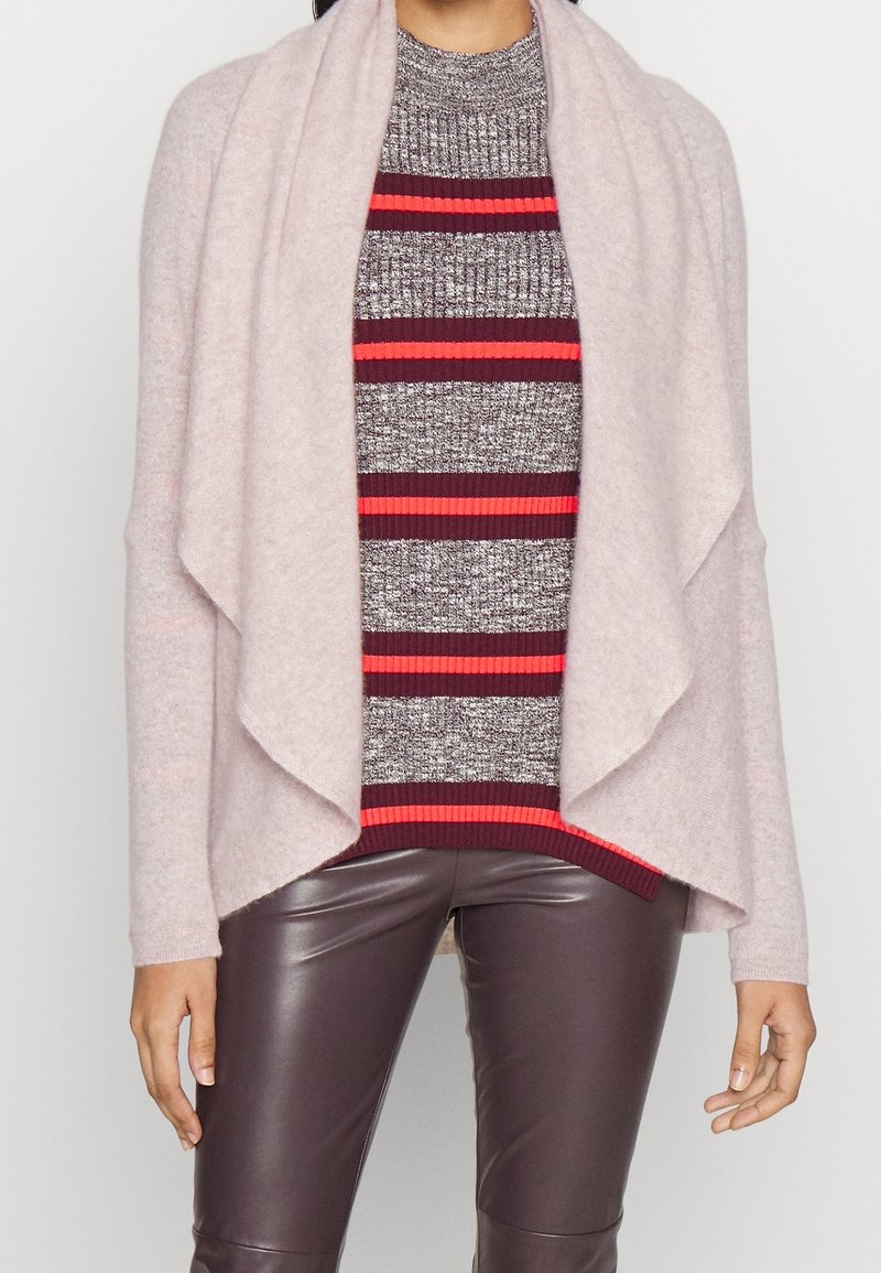 Helles rosa Cardigan mit drapierter Front, getragen über einem grau-rot gestreiften Pullover, kombiniert mit braunen Kunstlederhosen, die gerippte Akzente aufweisen.