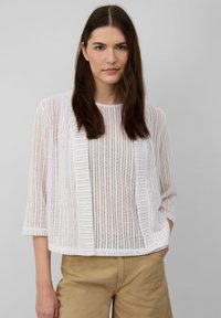 Witte, lichte gebreide trui met verticale strepen, open voorkant, driekwart mouwen en een relaxte pasvorm. Gedragen over tan kleurige broeken.