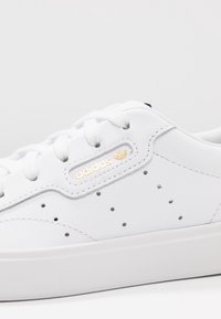 Vit lädersneaker med perforerade detaljer, grå accenter och en gyllene "adidas"-logotyp på sidan, med vita snören och en texturerad sula.