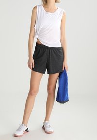 Žena v bílém vršku bez rukávů a tmavě šedých šortkách držící modrý ručník, obutá v bílých teniskách, stojící na pozadí bez vzoru.