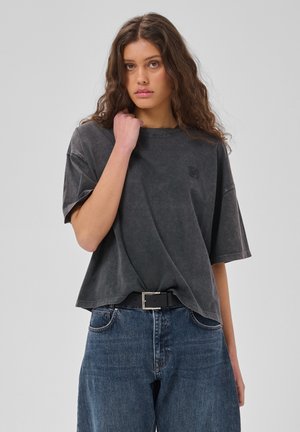 Kvinde med langt bølget hår har en løs mørkegrå T-shirt, blå jeans og et sort bælte, stående mod en ensfarvet lys baggrund.