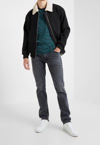 Hombre vestido con una chaqueta negra con cuello de forro polar blanco, una camisa a rayas en tonos verde azulado y negro, jeans grises y zapatillas blancas, posando con la mano en el bolsillo.