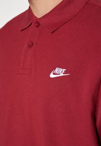 Polo rouge en tissu texturé, avec un col à deux boutons et un logo Nike brodé en blanc sur la poitrine gauche.