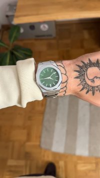 Ceas de mână din metal cu carcasă octogonală argintie, cadran texturat verde, marcaje orare argintii și brățară din oțel inoxidabil, purtat pe un încheietură cu un tatuaj.