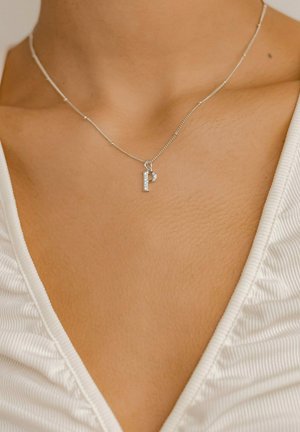 Silberne Halskette mit einem dekorativen "P" Anhänger, der mit einem kleinen Strassstein verziert ist und auf einem glatten, nackten Schlüsselbein ruht.