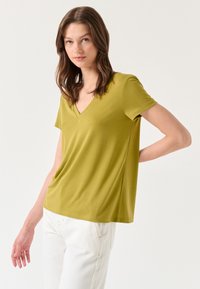 Olijfgroene V-hals T-shirt met korte mouwen, gemaakt van zachte stof. Heeft een ontspannen pasvorm en zijopeningen voor comfort. Gecombineerd met witte broek.