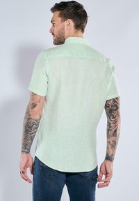 Helles mintgrünes kurzärmliges Hemd mit Kragen, das eine subtile Textur und abgerundeten Saum aufweist, getragen über dunkelblauen Jeans.