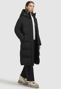 Lang sort pufferjakke med høj krave og lynlås foran. Har store quiltede sektioner, lommer, og er parret med beige chunky støvler.