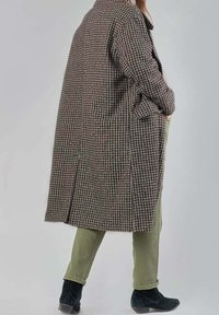 Abrigo largo de cuadro en marrón y beige con cuello, bolsillos laterales y un corte holgado, usado sobre pantalones verde oliva y botas negras de tobillo.