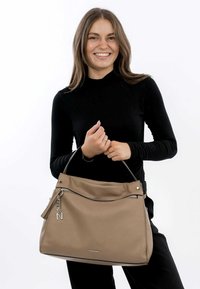 Beige synthetic tote bag with a zippered front pocket, silver hardware accent, and a curved top. Model holds the bag with a slim handle.