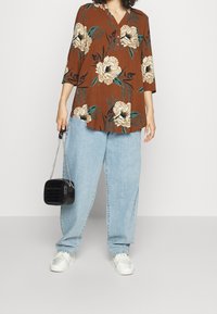 Tunika floreale marrone con scollatura a bottoni, abbinata a jeanslarghi blu chiaro. Tiene in mano una piccola borsa nera in finta pelle di coccodrillo, indossando sneakers bianche.